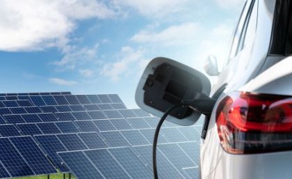 The side of an electric car, plugged in to a charging outlet with solar panels in the background.